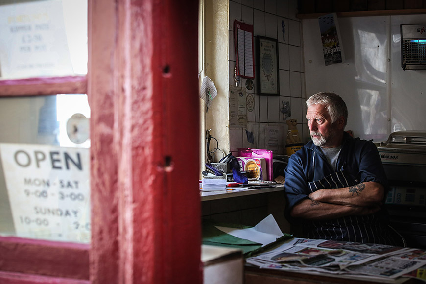 Man sat down behind shop counter gazing outside by Ceri Oakes