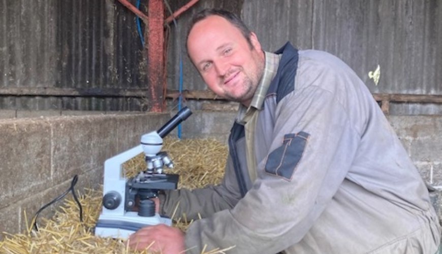 A person stands in a barn, leaning over a hay bale which holds a microscope. The person is looking directly at the camera and smiling.