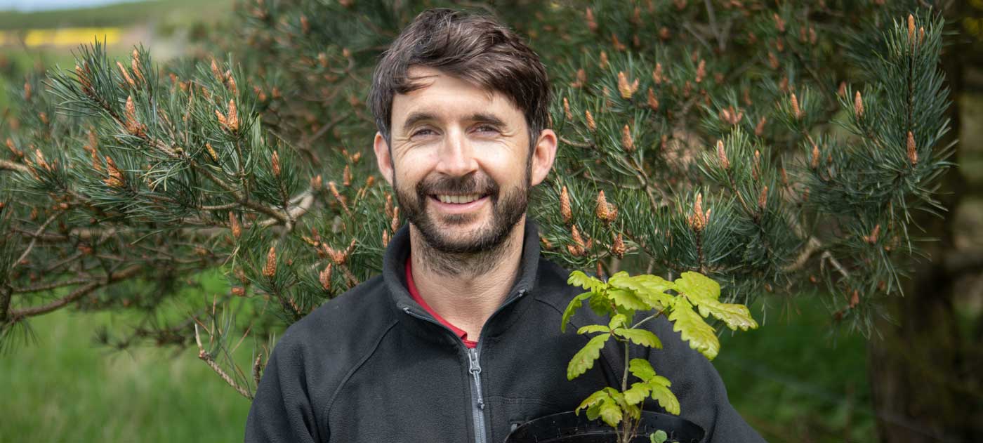Man holding an oak sapling in front of a tree. Credit Charlie Fox. Man holding an oak sapling in front of a tree. Credit Charlie Fox.