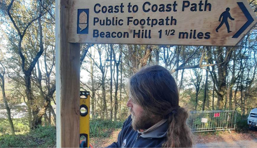 Person standing by a wooden signpost for a public footpath in a wooded area.