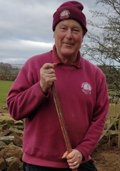 Paul Welford stands in branded North York Moors clothing holding what could be a rake or long tool