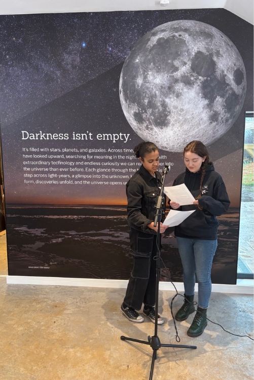 Two individuals stand together at a microphone in front of a wall featuring a large moon and a starry sky, sharing a document.