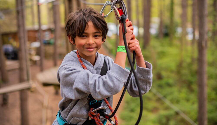 A young boy is in a harness at Go Ape, smiling and looking at the camera.