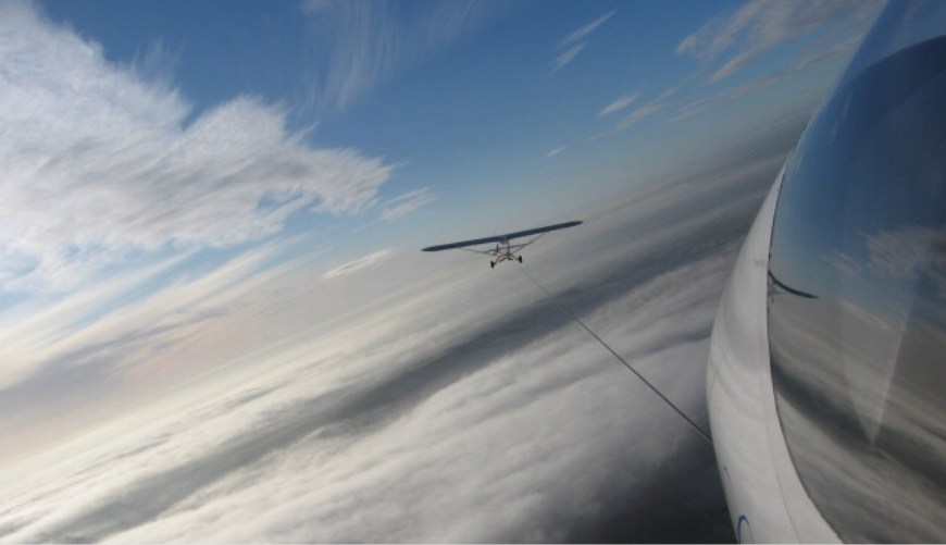 A small plane tows a glider above clouds with a clear blue sky in the background.