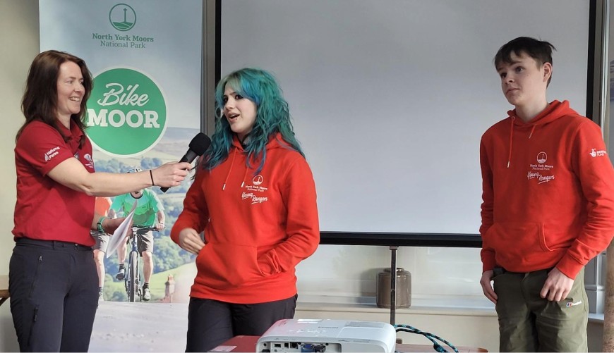 Three people stand in front of a screen; one holds a microphone, speaking to the others.