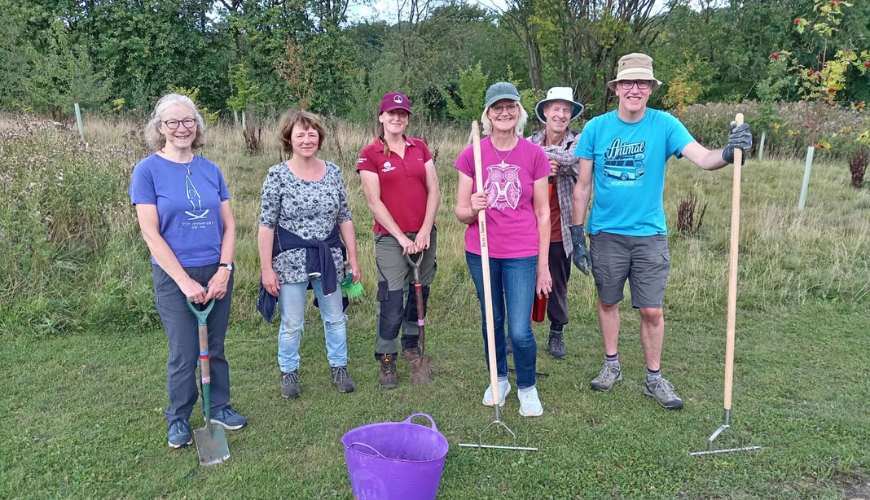 A group of six smiling people stand on grass holding gardening tools, with trees in the background, conveying teamwork and community spirit.