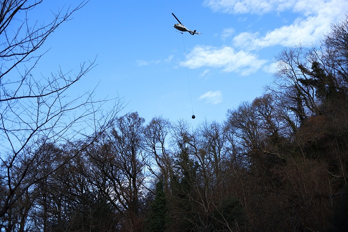 Helicopter flying in blue sky above trees, hanging some distance below it is a bag of stone