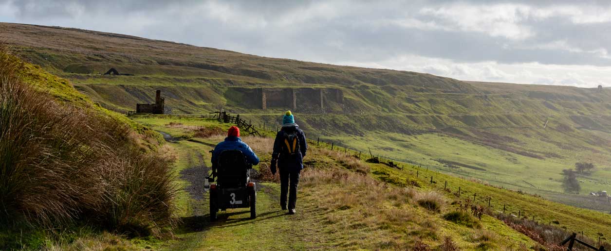 Two people heading down an old railway track towards industrial monuments in the distance. One person is using a all-terrain mobility scooter and the other is walking alongside. Two people heading down an old railway track towards industrial monuments in the distance. One person is using a all-terrain mobility scooter and the other is walking alongside.