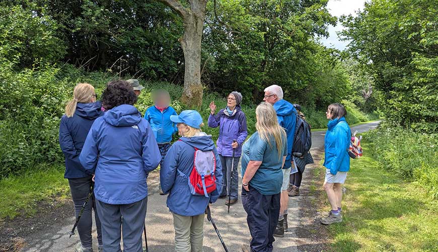 group of people stood in a circle outside listening to someone speak as they point at the trees