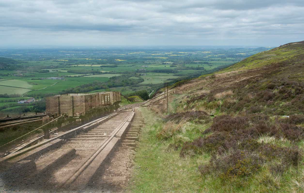 Photo showing a steep slope heading down into a farmed and wooded landscape. There is another image which has been edited on showing an industrial railway heading down the slope. Credit John Davies of the Kirby, Great Broughton and Ingleby Greenhow Local History Group. Photo showing a steep slope heading down into a farmed and wooded landscape. There is another image which has been edited on showing an industrial railway heading down the slope. Credit John Davies of the Kirby, Great Broughton and Ingleby Greenhow Local History Group.