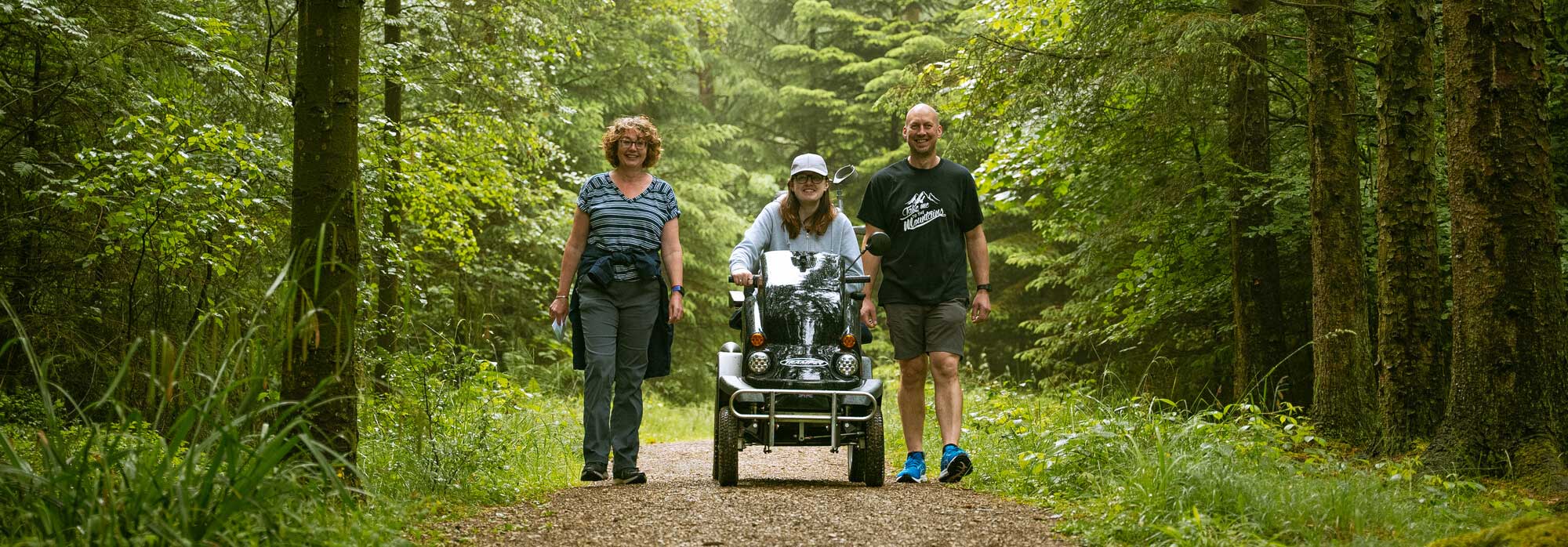 Three people heading down a woodland track. Two people are walking either side of another person using a tramper (all-terrain mobility scooter). Credit Hewitt & Walker.