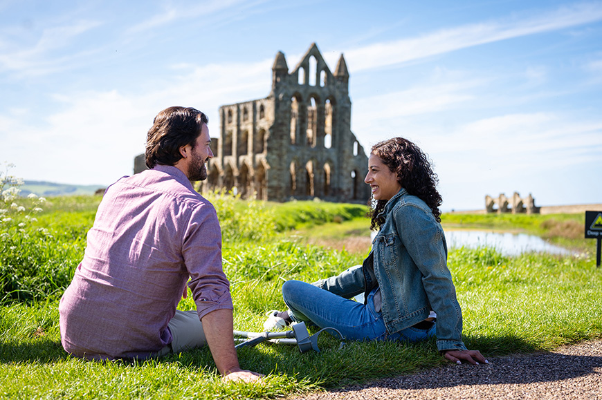 Man and woman sitting on grass with crutches beside them, Whitby Abbey in the background Credit VisitBritain/Peter Kindersley