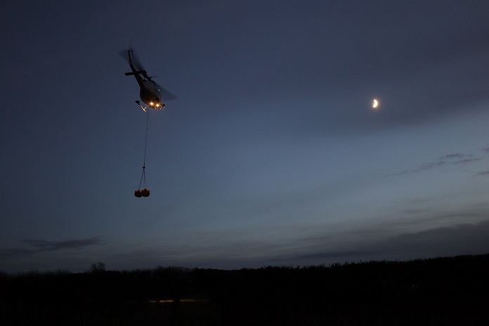 A clear night sky with the moon visible. A helicopter flies to the left of the picture and suspended below it our two bags.  
