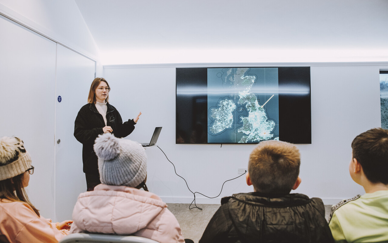 A woman presenting in the Dark Skies Station. The room is white with an audience sat in front of a television with the UK map and a laptop.