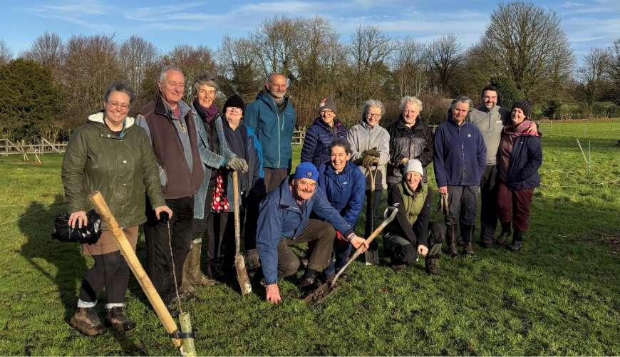 A group of people stand in a field, they are wearing coats, hats and gloves and holding spades. It is a cold but sunny day.