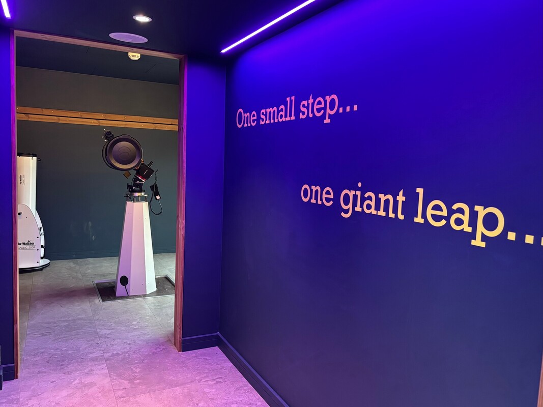 A blue painted wall with the words 'one small step... one giant leap...'. The corridor leads to a room with an open door showing a large telescope.