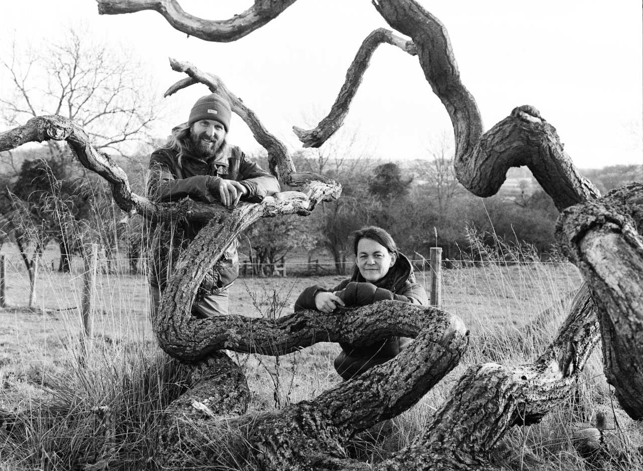 Rosey and Fraser. Two people standing among a fallen tree in a field.