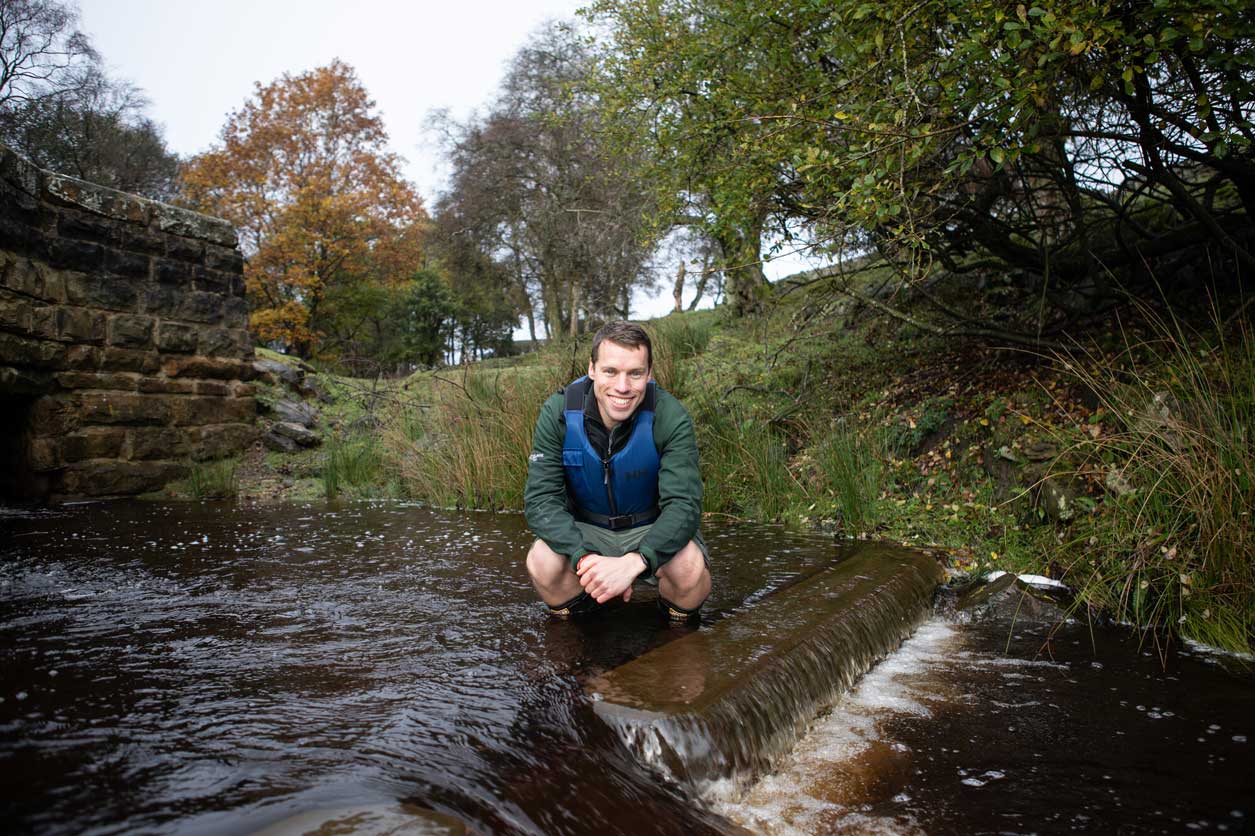 Male posing for a photo in a river. He is psotioned next to a wooden pre-weir, which has been installed as part of efforts to improve fish passage through the water. Credit Charlie Fox.