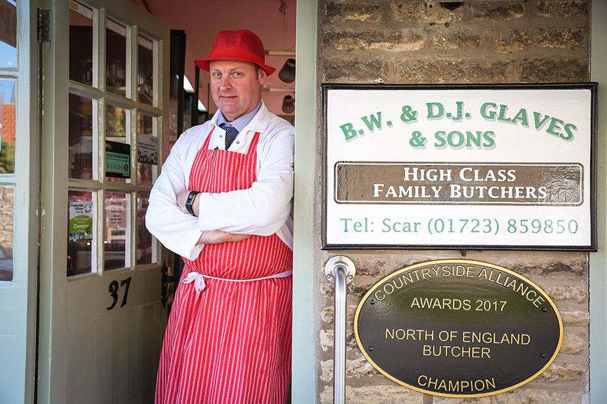 Butcher stood in the front door beside sign and award plaque by Ceri Oakes