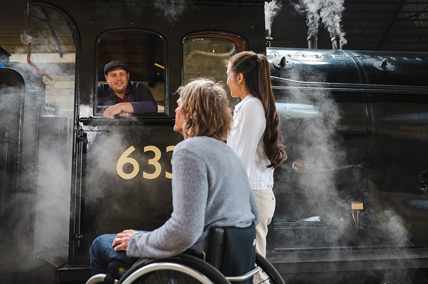 Man in a wheelchair and woman talking to the driver of a steam engine No 63395, surrounded by steam Credit VisitBritain-Peter Kindersley