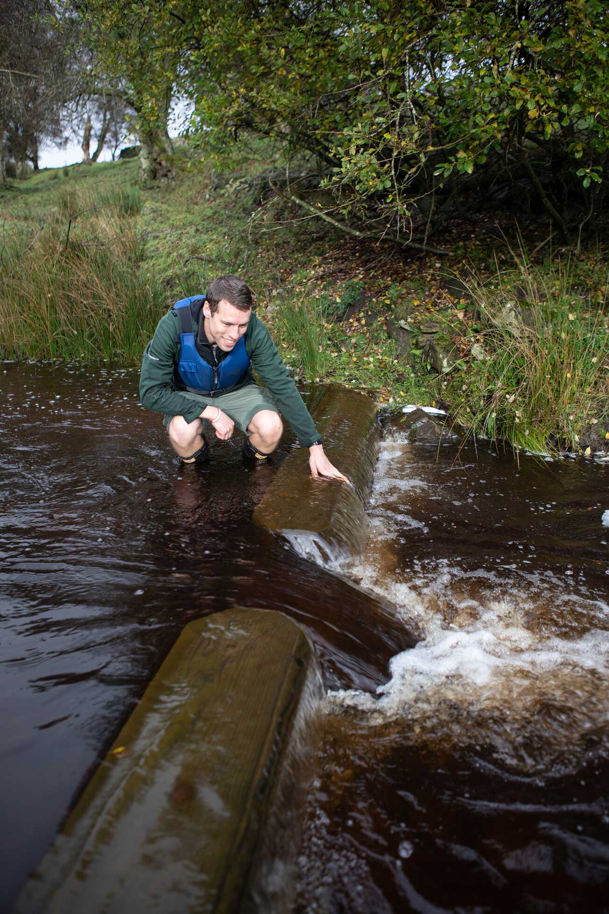 Male next to a wooden pre-weir in a river. The pre-weir has been installed as part of efforts to improve fish passage through the water. Credit Charlie Fox.