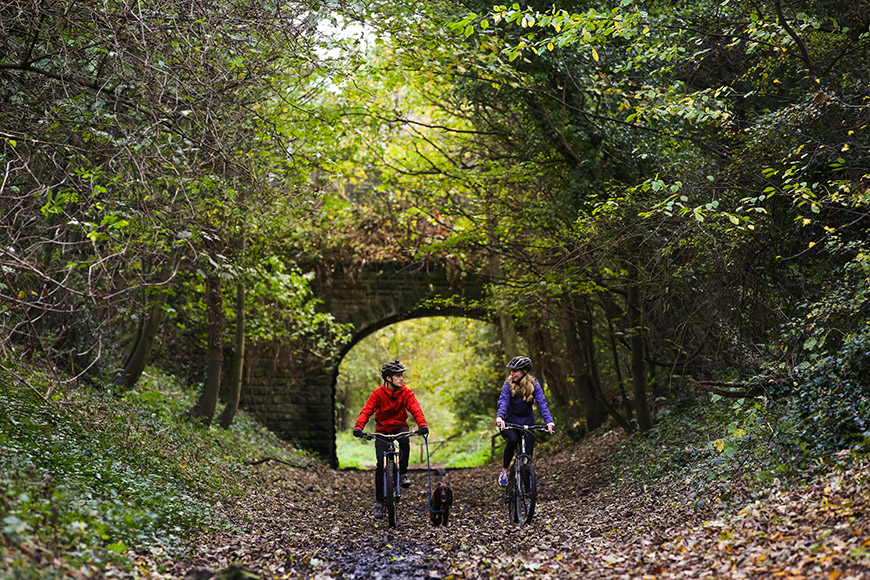 Two people biking on the Cinder Track with a dog on a lead