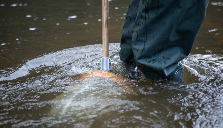 A person in green waders stands in shallow, rippling water holding a wooden pole.