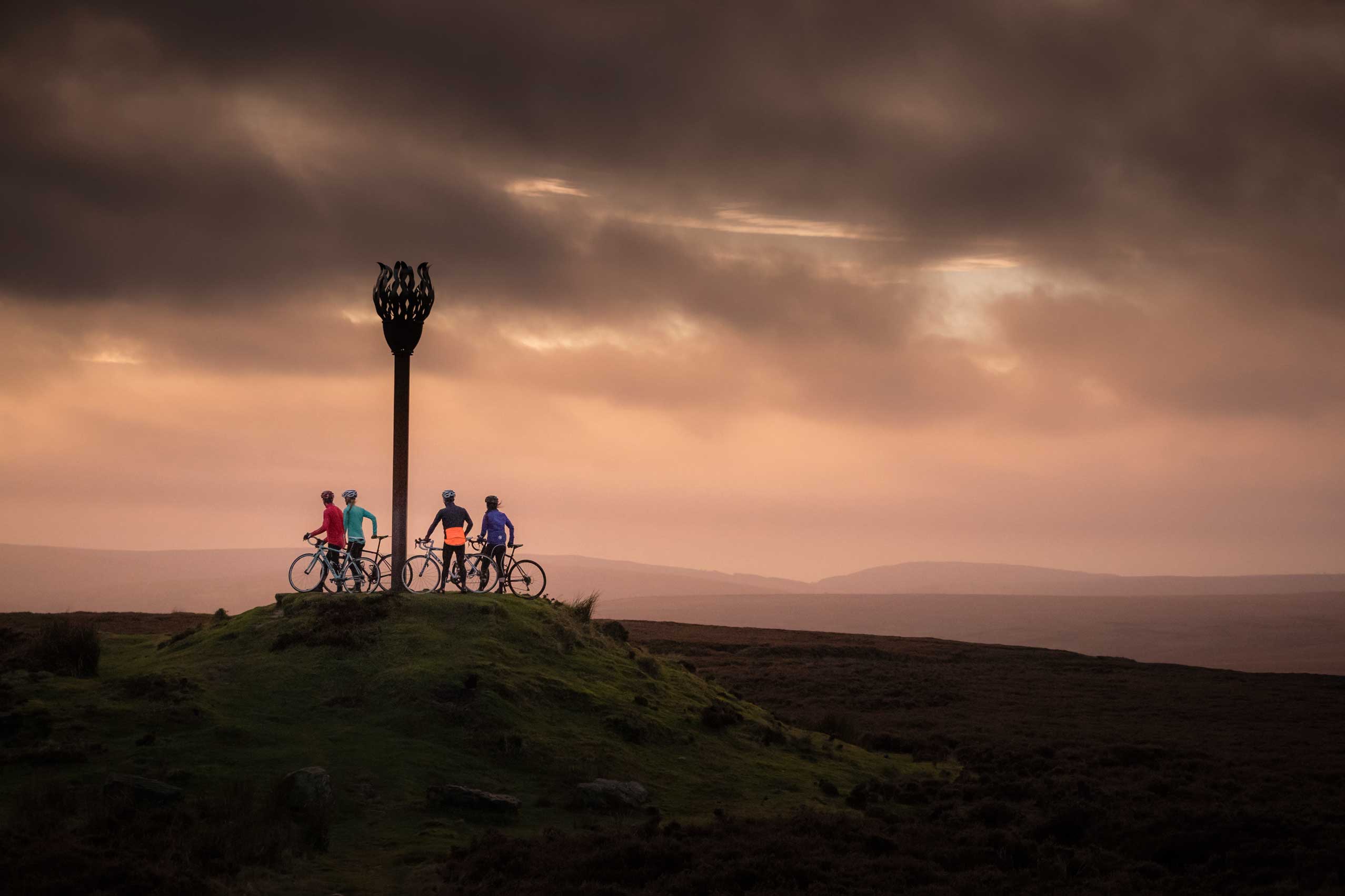 Cyclists stood beneath Danby Beacon with dramatic clouds in the distance. Credit Russell Burton/NYMNPA.