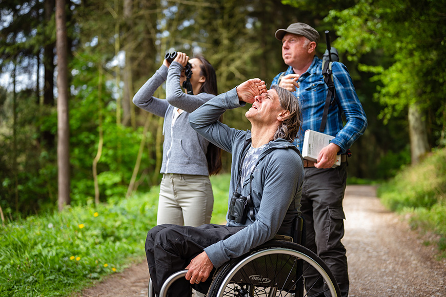 Man in checked shirt with telescope is telling two people, a woman using binoculars and a man using a wheelchair about the forest bird song Credit VisitBritain/Peter Kindersley