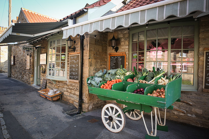 Exterior of butchers with a green cart containing vegetables by Ceri Oakes