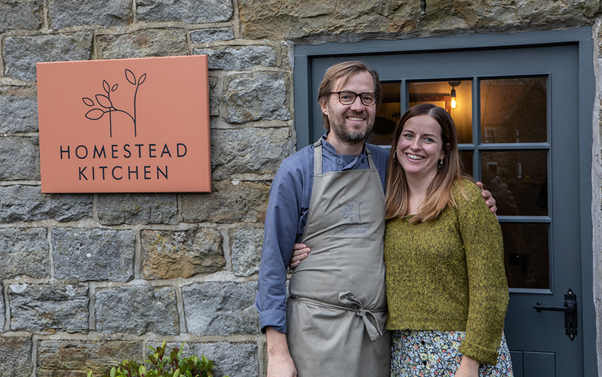 Peter and Cecily stood outside front door beside Homestead Kitchen sign by Polly Baldwin