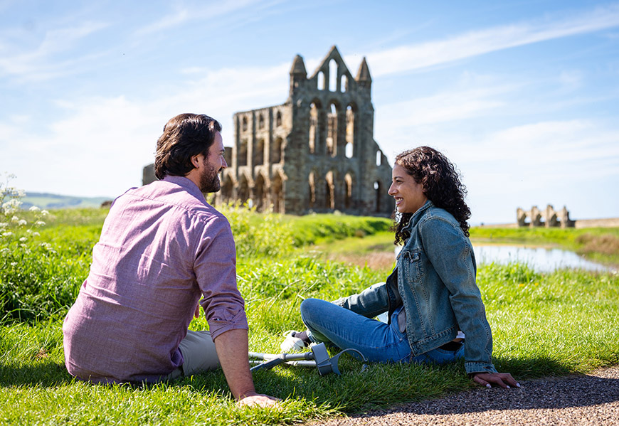 Man and woman sitting on grass with crutches beside them, Whitby Abbey in the background Credit Visit Britain/Peter Kindersley