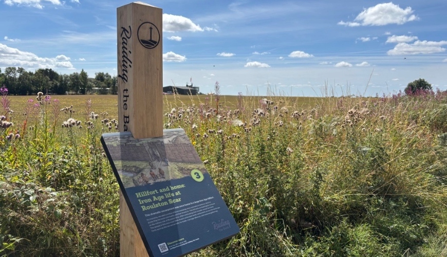 Hood Hill information panel on the Raiding the Bank trail A wooden signpost in a scenic landscape displays a castle illustration and text about Hood Hill, with green hills and a blue sky in the background.