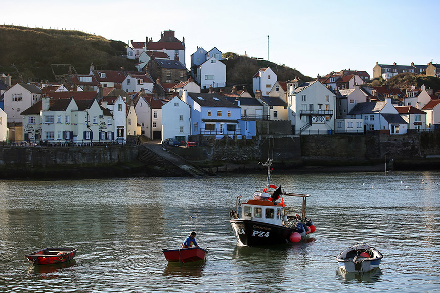 Staithes harbour with boats in the water and village in the background by Ceri Oakes