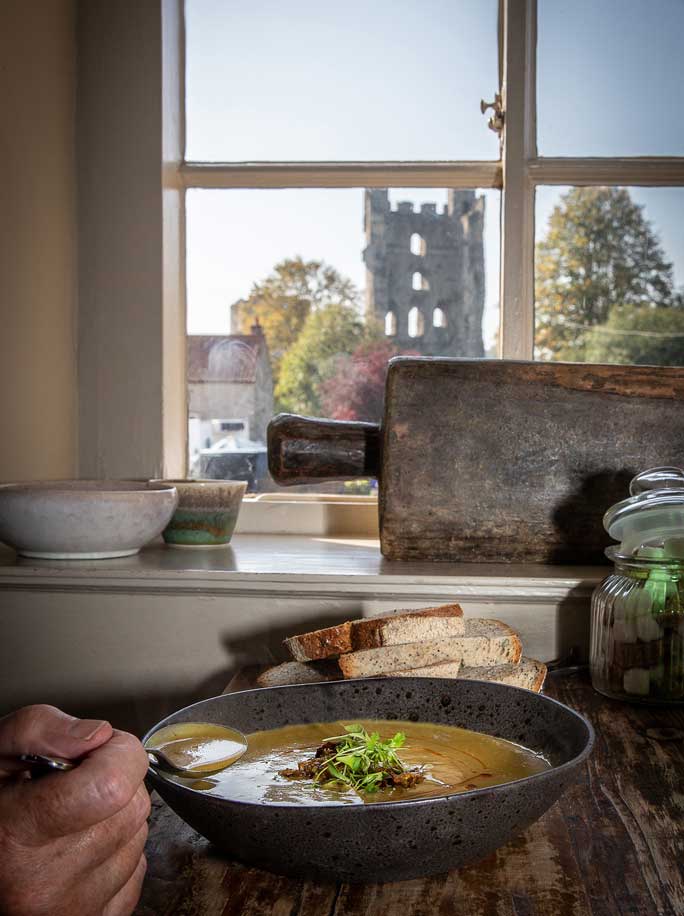 Bowl of soup on table with Helmsley Castle in the view outside by Polly Baldwin
