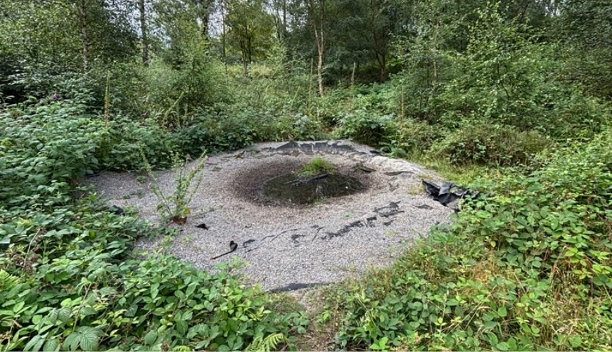 Circular gravel depression in a forest surrounded by dense green vegetation, with black material along the edges.