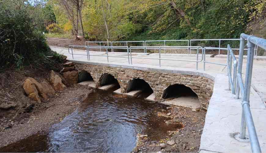 Stone bridge with four arches over a shallow stream, surrounded by green foliage. The concrete pathway has metal railings, conveying a calm, natural setting.