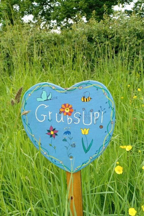 Blue heart-shaped sign in a meadow with "Grub's UP!" painted on it, surrounded by illustrations of flowers and insects.