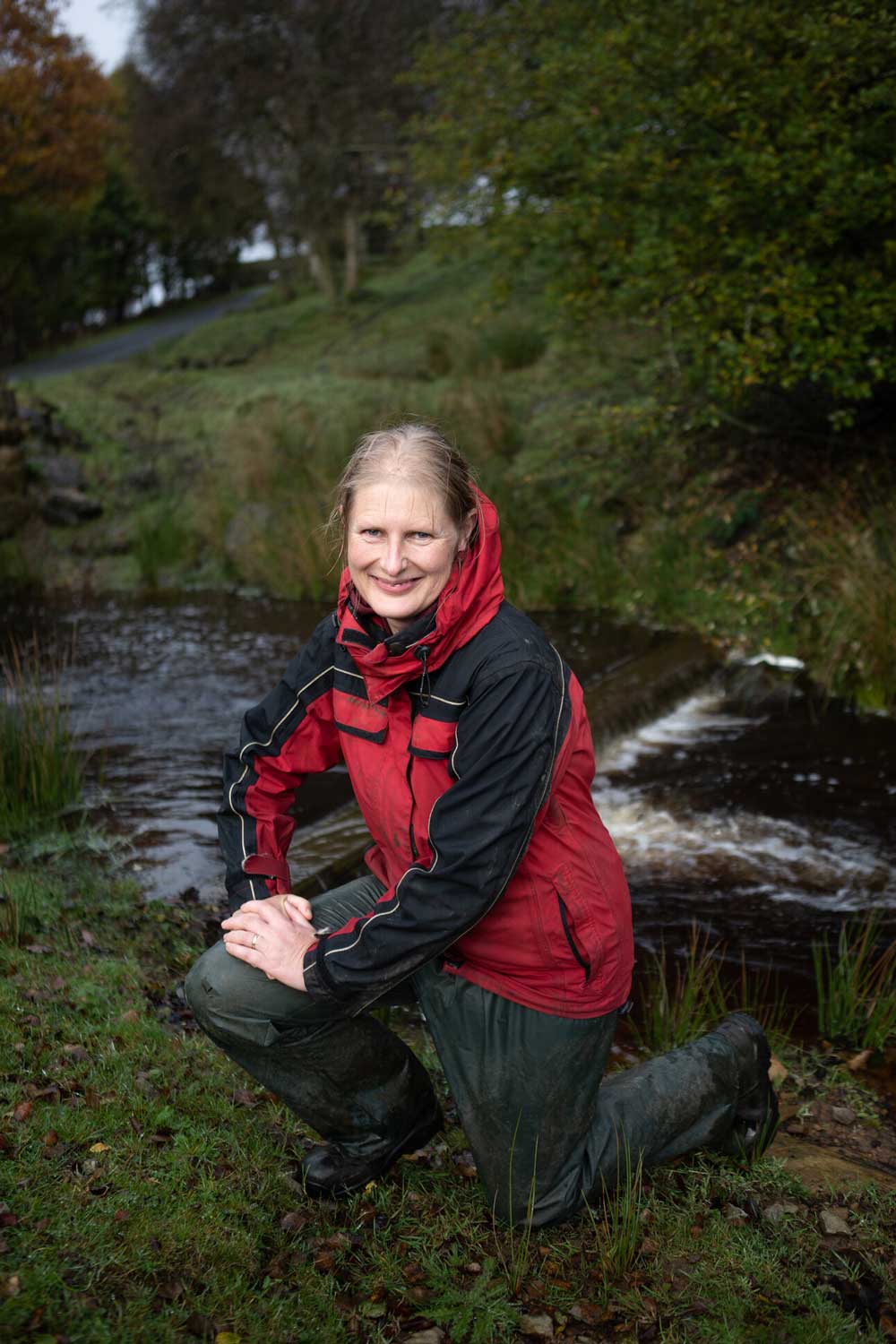 Trudy Sanderson (upland livestock farmers) kneeling in front of a stone bridge and river. Credit Charlie Fox.